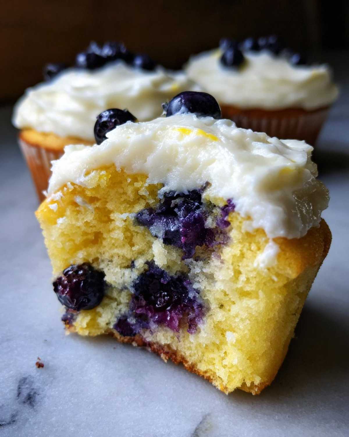 Close-up of a Lemon Blueberry Cupcakes with Cream Cheese Frosting cut in half, showing moist yellow cake and burst blueberries.