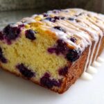 A close-up of a freshly baked Lemon Blueberry Yogurt Loaf showing the moist yellow crumb studded with blueberries and topped with a white lemon glaze.