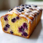 A close-up of a moist Lemon Blueberry Yogurt Loaf, featuring bright yellow cake studded with blueberries and drizzled with white lemon glaze.