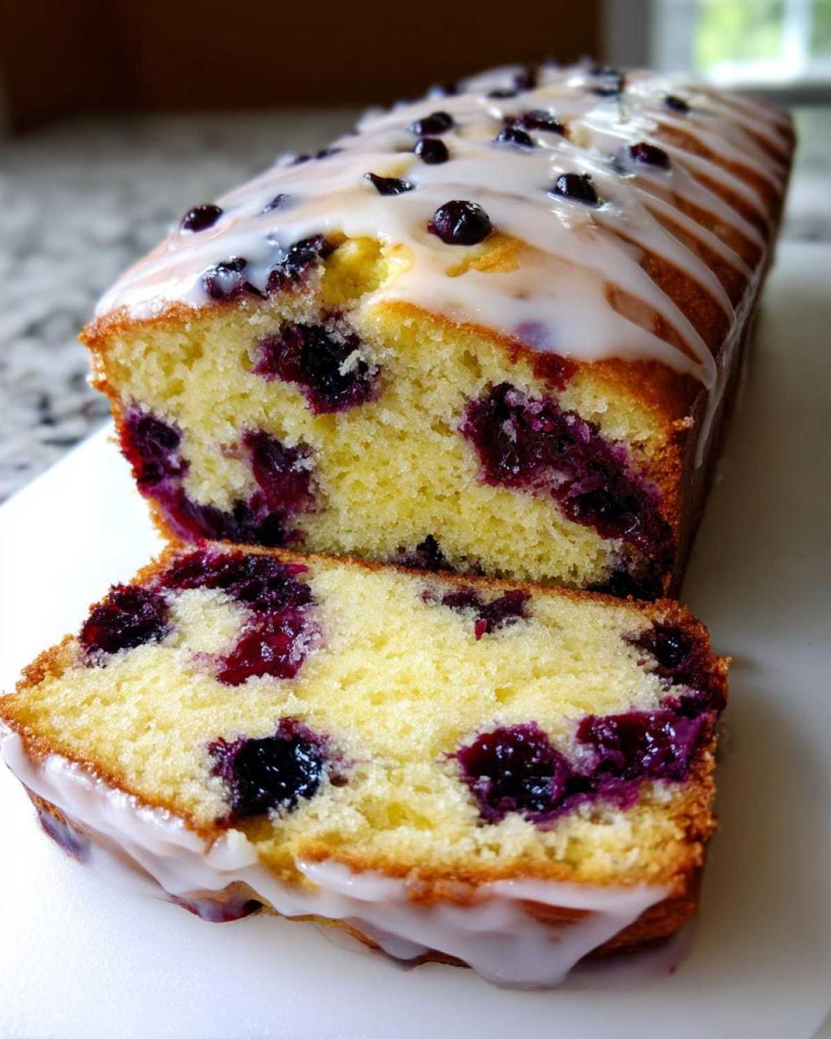 A close-up of a sliced Lemon Blueberry Yogurt Loaf showing moist crumb and bursting blueberries, topped with a lemon glaze.
