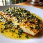 Close-up of a perfectly cooked Lemon Garlic Butter Tilapia fillet swimming in bright yellow butter sauce with chopped parsley and garlic.