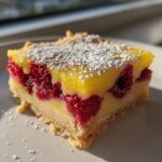 A single square of Lemon Raspberry Bars featuring a bright yellow lemon layer, whole raspberries, and a shortbread crust, dusted with powdered sugar.