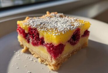 A single square of Lemon Raspberry Bars featuring a bright yellow lemon layer, whole raspberries, and a shortbread crust, dusted with powdered sugar.