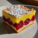 A square slice of Lemon Raspberry Bars featuring a shortbread crust, a layer of bright red raspberries, and a thick lemon topping dusted with powdered sugar.