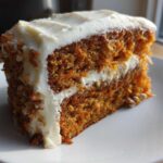 A close-up of a thick slice of Moist Carrot Cake with Cream Cheese Frosting, showing visible shredded carrots.
