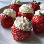 Close-up of several No-Bake Cheesecake Stuffed Strawberries topped with swirls of white whipped cream on a white plate.