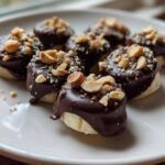 Close-up of several No-Bake Chocolate Covered Banana Bites topped with chopped peanuts on a white plate.