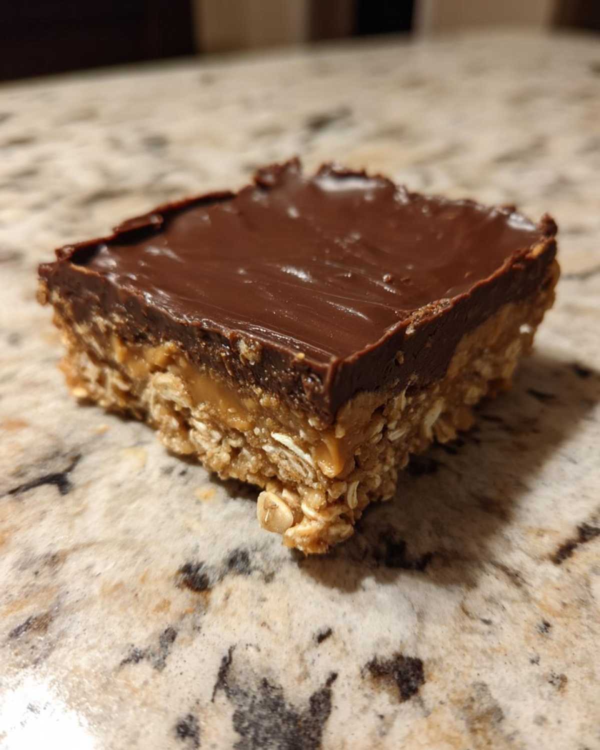 A single square of No-Bake Chocolate Peanut Butter Oat Bars with a thick chocolate topping resting on a granite counter.