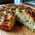 A piece of golden-brown No-Knead Skillet Focaccia with Rosemary, showing its airy interior texture, topped with herbs and sea salt.