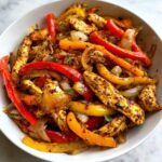 Close-up of seasoned chicken strips mixed with sautéed red, yellow, and orange bell peppers and onions in a white bowl.