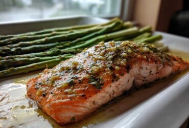 A perfectly cooked fillet of One-Pan Lemon Garlic Salmon topped with herbs, next to roasted asparagus spears.