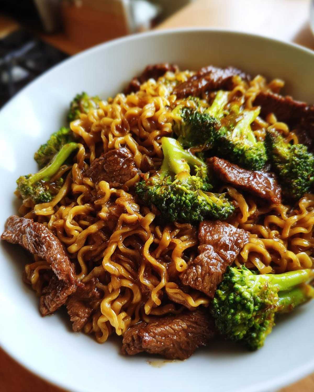 A close-up of One-Pot Beef and Broccoli Ramen Noodles coated in savory sauce, served in a white bowl.