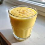 A thick, bright yellow Pineapple Coconut Smoothie topped with toasted coconut flakes in a clear glass.