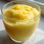 A close-up of a thick, bright yellow Pineapple Coconut Smoothie served in a clear glass, topped with small fruit pieces.