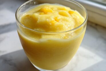 A close-up of a thick, bright yellow Pineapple Coconut Smoothie served in a clear glass, topped with small fruit pieces.