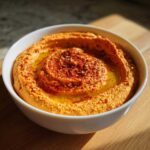 A close-up of creamy Roasted Red Pepper Hummus swirled in a white bowl, topped with olive oil and paprika.