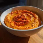 A close-up of creamy Roasted Red Pepper Hummus swirled with olive oil and sprinkled with paprika in a white bowl.