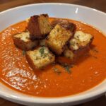 A bowl of vibrant orange Roasted Tomato Basil Soup topped with golden brown Grilled Cheese Croutons and dried herbs.