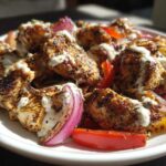 Close-up of seasoned Sheet Pan Chicken Shawarma pieces drizzled with garlic yogurt sauce and served with red onion and bell peppers.