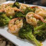 Cooked shrimp and roasted broccoli florets coated in garlic butter sauce on a white platter.
