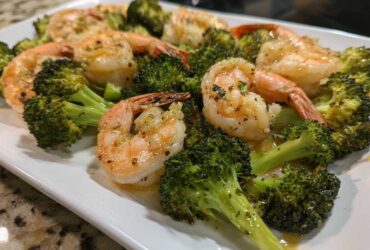Cooked shrimp and roasted broccoli florets coated in garlic butter sauce on a white platter.