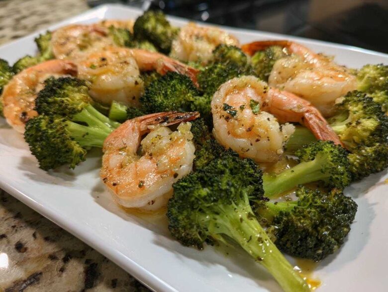 Cooked shrimp and roasted broccoli florets coated in garlic butter sauce on a white platter.