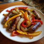 A white plate filled with browned sausages, caramelized onions, and colorful roasted bell peppers from the Sheet Pan Sausage and Peppers recipe.