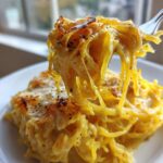Close-up of cheesy Spaghetti Squash Alfredo strands being lifted high on a fork from a serving on a white plate.
