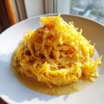 A close-up of bright yellow Spaghetti Squash Alfredo piled high on a white plate with creamy sauce.