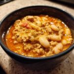 A close-up of a dark bowl filled with hearty Spicy Turkey White Bean Chili, showing ground turkey, white beans, and a rich orange broth.