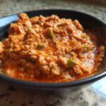Close-up of a rich, orange-red serving of Spicy Turkey White Bean Chili with visible ground turkey and green peppers in a dark bowl.