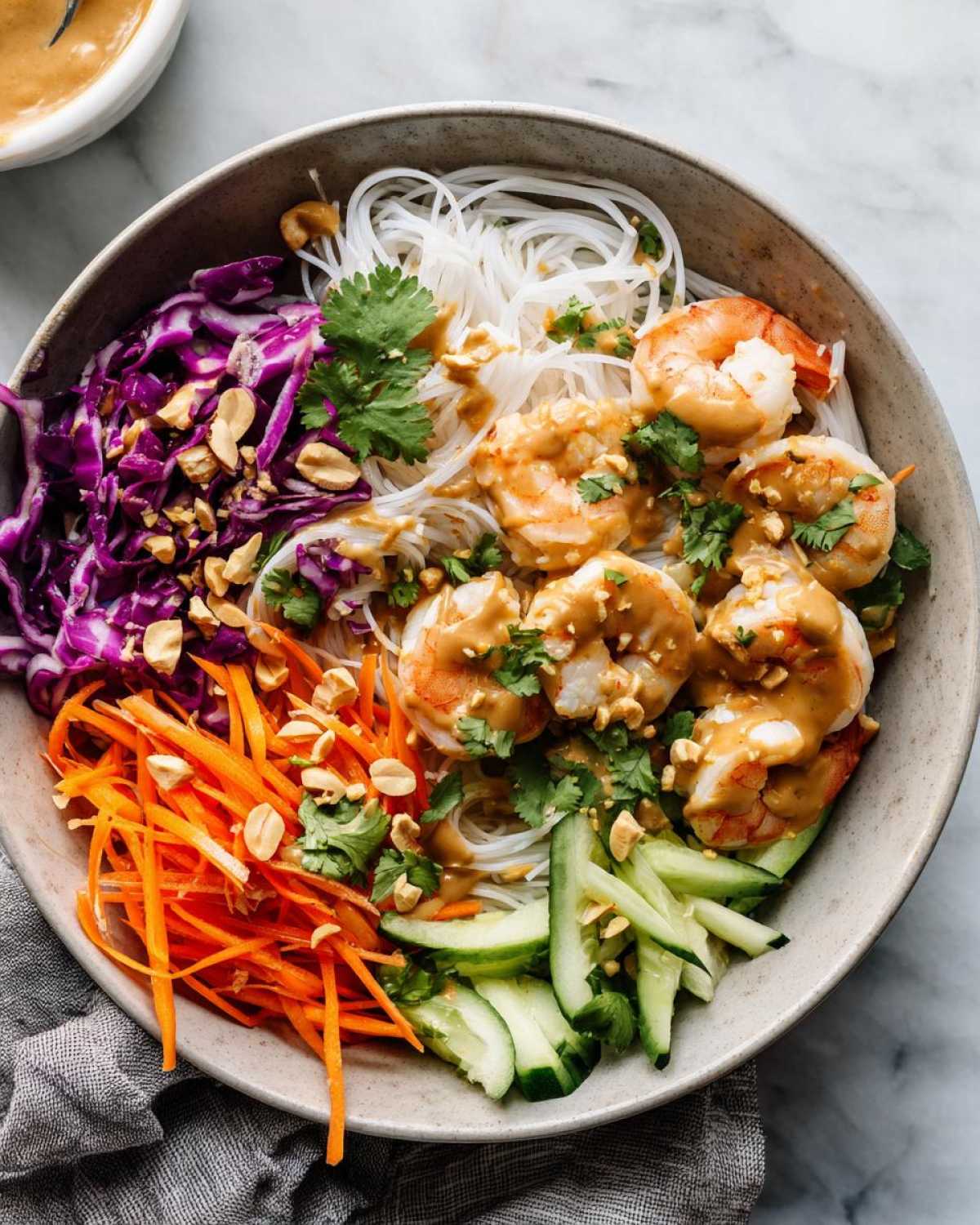 Overhead view of a colorful Spring Roll Bowl with Peanut Sauce, featuring shrimp, rice noodles, carrots, cucumber, and red cabbage.