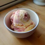 Two scoops of swirled Strawberry Banana Ice Cream in a white bowl on a wooden surface.