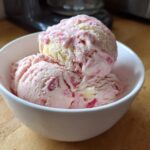 Two scoops of pink and white Strawberry Banana Ice Cream served in a small white bowl on a wooden surface.