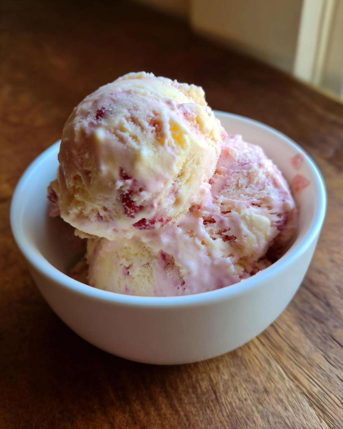 Two scoops of creamy, swirled Strawberry Banana Ice Cream served in a small white bowl on a wooden surface.