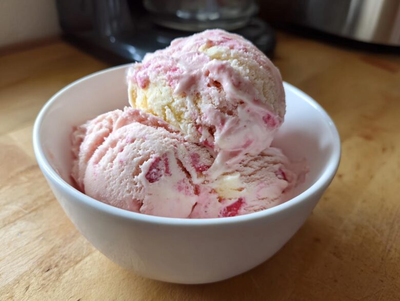 Two scoops of pink and white Strawberry Banana Ice Cream served in a small white bowl on a wooden surface.