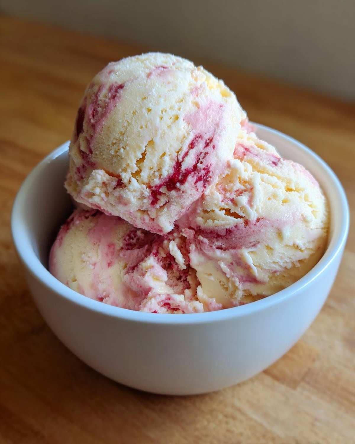 Three scoops of creamy Strawberry Banana Ice Cream with visible swirls of bright pink fruit puree in a white bowl.