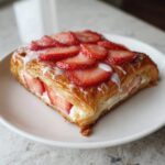 A flaky, golden Strawberry Cream Cheese Danish topped with sliced strawberries and white icing, served on a white plate.