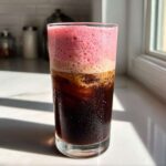 A tall glass of Strawberry Cream Cold Brew showing dark coffee below and thick pink foam on top.