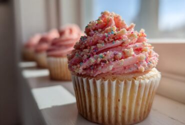 A close-up of a single Strawberry Crunch Cupcakes featuring pink frosting topped with colorful sprinkles and crunch pieces.