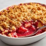 A close-up of a spoonful being taken from a warm Strawberry Rhubarb Crisp, showing bubbling red fruit filling and golden crumble topping.