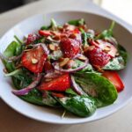 A close-up of a vibrant Strawberry Spinach Salad with poppy seed dressing, sliced strawberries, red onion, and slivered almonds.