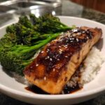 A close-up of a Teriyaki Salmon Rice Bowls serving featuring a glazed salmon fillet over white rice and bright green broccoli.