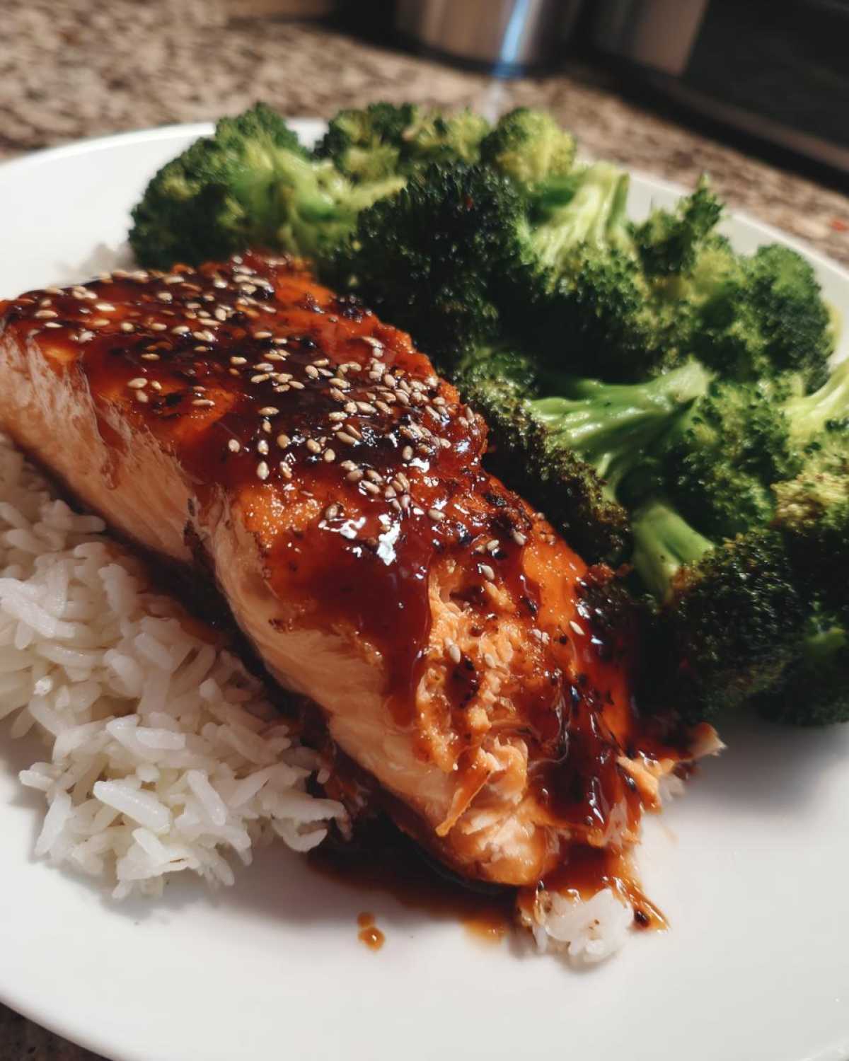 Close-up of a flaky salmon fillet covered in thick teriyaki glaze and sesame seeds, served over white rice with steamed broccoli.