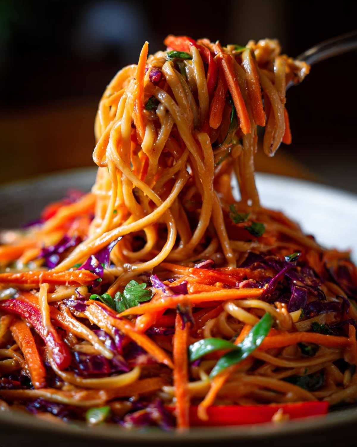A fork lifts a generous serving of saucy Thai Peanut Noodles with Crunchy Veggies, featuring bright orange carrots and purple cabbage.
