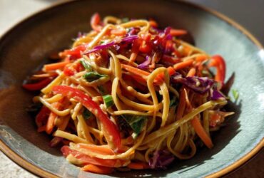 Close-up of a bowl of Thai Peanut Noodles with Crunchy Veggies, featuring noodles coated in peanut sauce and mixed with bright red peppers and carrots.