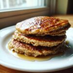 A close-up stack of three golden-brown Vegan Banana Oat Pancakes drizzled with syrup.