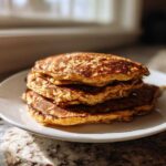 A stack of four golden-brown Vegan Banana Oat Pancakes sitting on a white plate near a sunlit window.