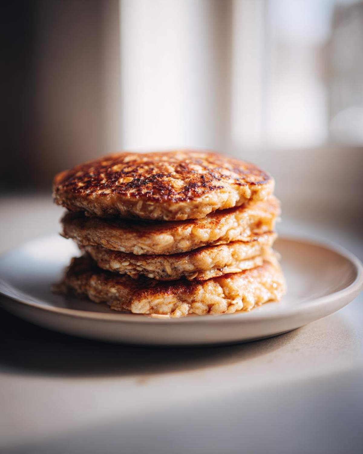 A close-up stack of four golden-brown Vegan Banana Oat Pancakes resting on a small, light-colored plate.