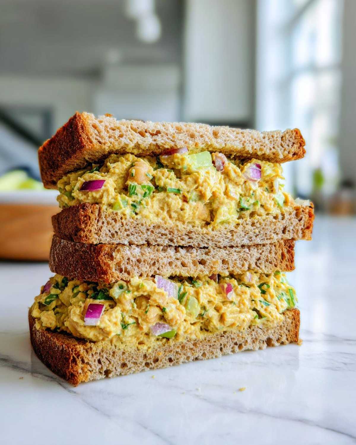 Two halves of a Vegan Chickpea Salad Sandwich stacked high on marble, showing creamy chickpea filling with red onion and celery.